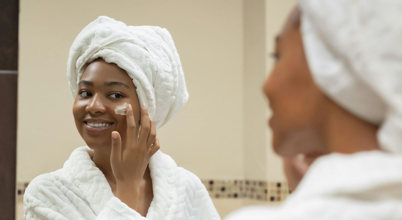 A smiling woman in a robe and head towel with cream on her cheek